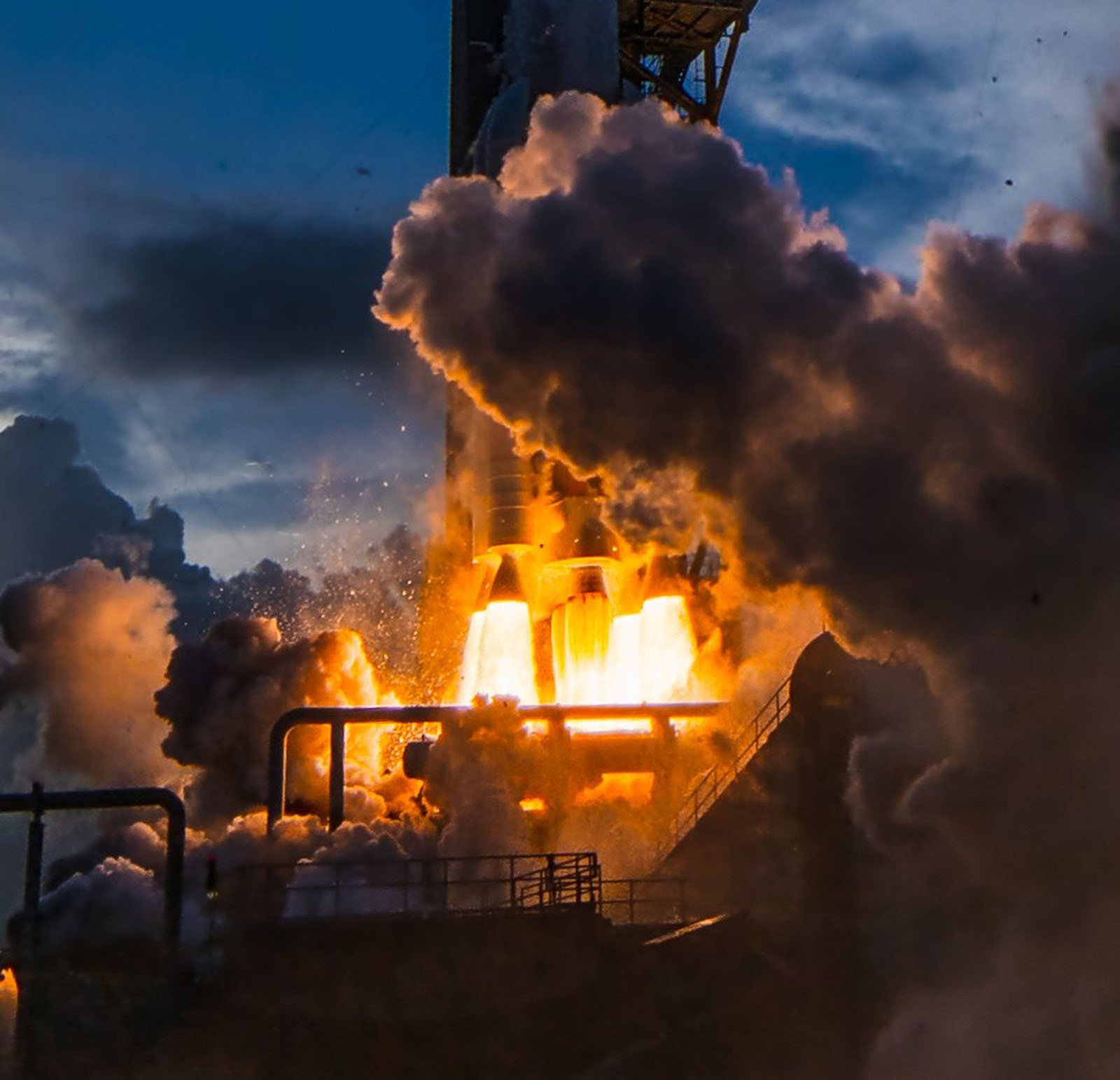 A close-up view of a rocket launch with bright flames and thick smoke billowing from the engines, illuminated against a dark, cloudy sky.
