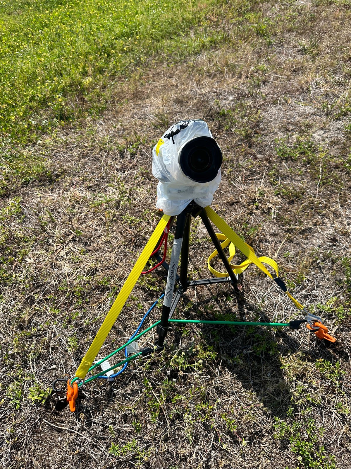 A camera covered in plastic is mounted on a tripod, which is secured with yellow straps and anchored to the ground with stakes and ropes in a grassy outdoor area.