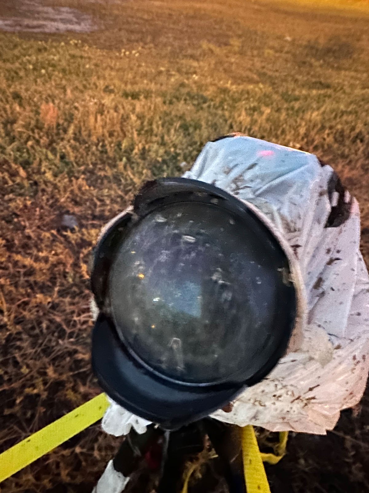 A covered surveying instrument, possibly a total station or camera, is mounted on a tripod in a grassy outdoor area at dusk. The lens appears dirty or fogged, and yellow tape is visible nearby.