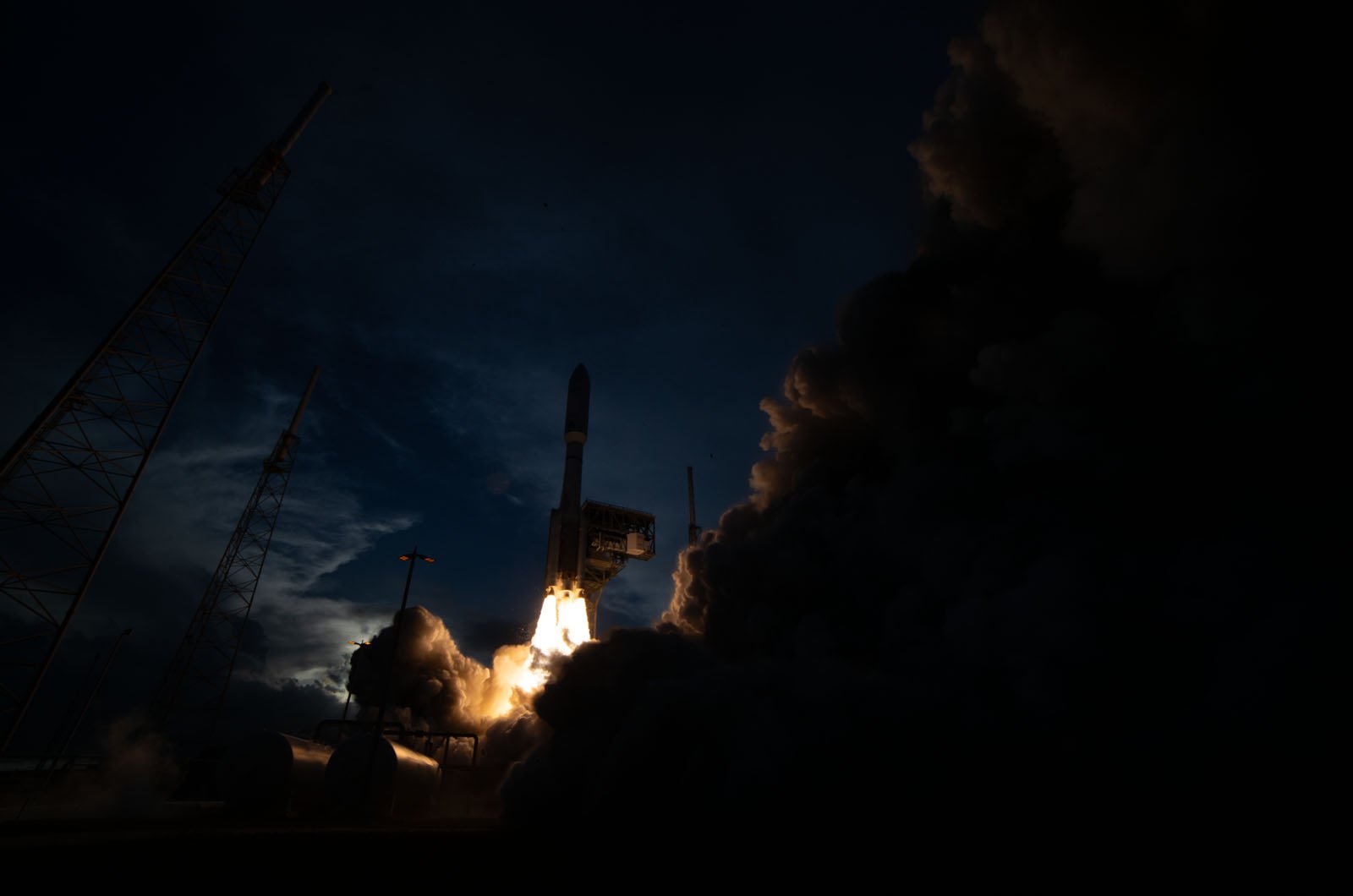 A rocket launches into a dark sky, illuminating the scene with bright flames and thick clouds of smoke, surrounded by tall support towers.