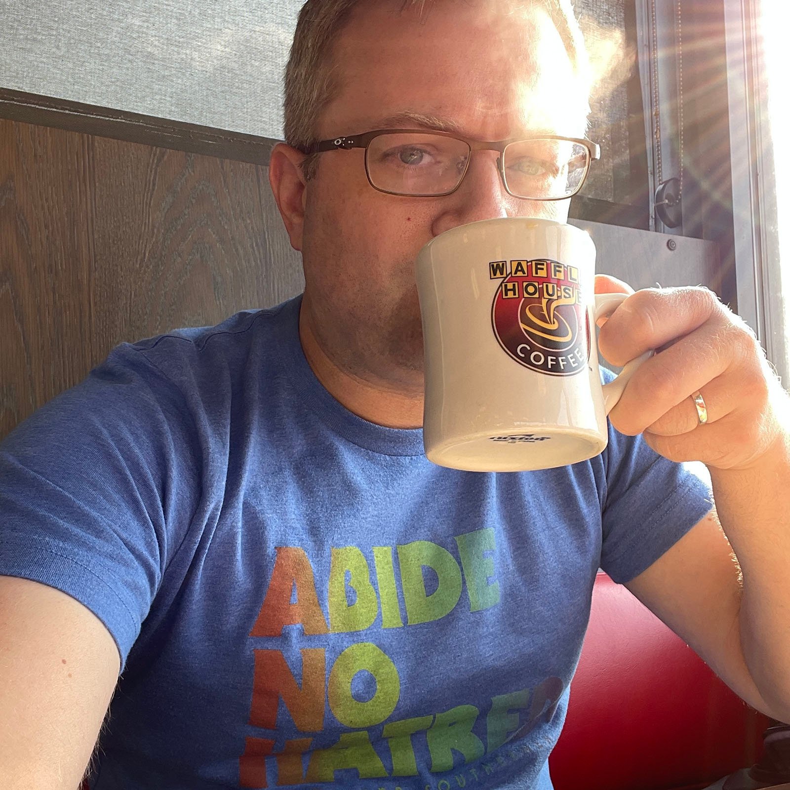 A person wearing glasses and a blue "Abide No Hatred" t-shirt sits in a sunlit booth, holding a Waffle House coffee mug and looking at the camera.