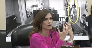 A woman in a pink top speaks at a meeting, gesturing with her hands. Behind her is a large black-and-white photo showing a living room and a figure highlighted in a yellow oval. A nameplate reads “Ms. Mace.”.