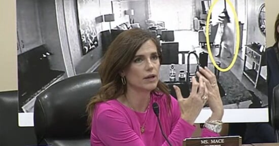 A woman in a pink top speaks at a meeting, gesturing with her hands. Behind her is a large black-and-white photo showing a living room and a figure highlighted in a yellow oval. A nameplate reads “Ms. Mace.”.
