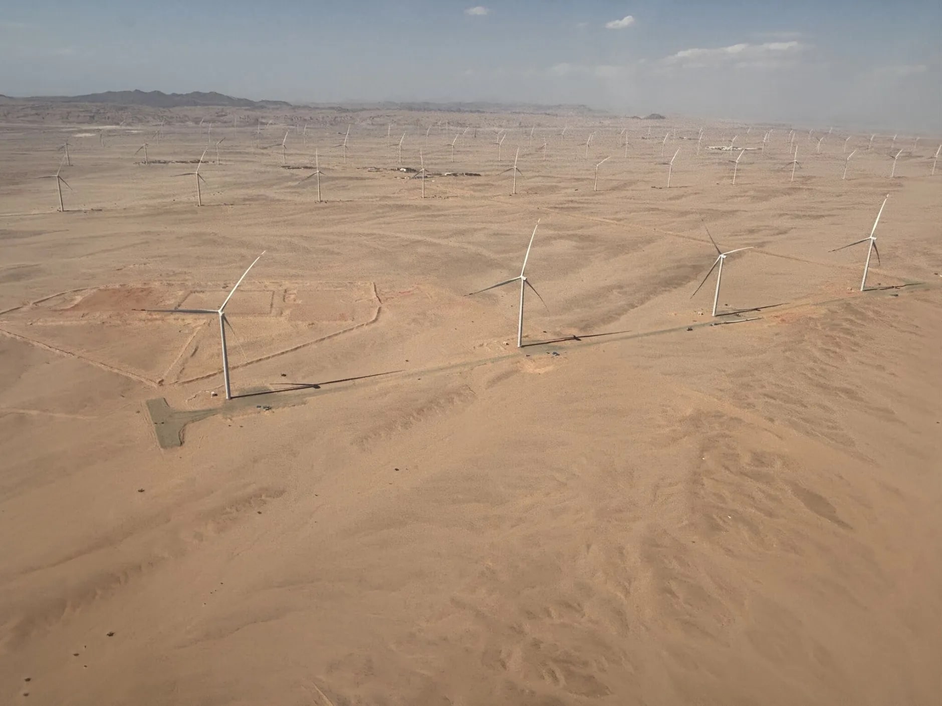 Aerial view of multiple wind turbines spread across a vast, sandy desert landscape under a clear sky. Mountains are visible on the horizon.