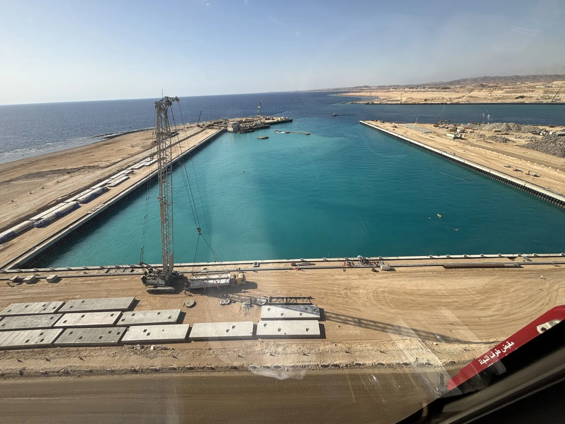 Aerial view of a construction site with a large, rectangular harbor surrounded by sandy land, a crane, construction materials, and the sea visible in the background under a clear sky.