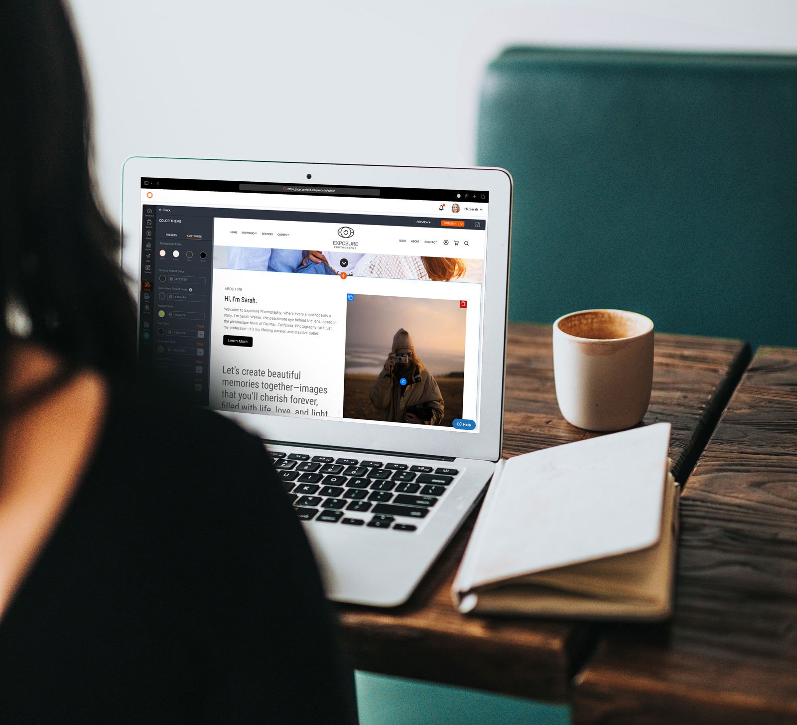 A person sits at a wooden table using a laptop, with an open notebook and a cup of coffee nearby. The laptop screen displays a website featuring images and text.