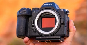 A close-up of a hand holding a Nikon Z series mirrorless camera with the lens removed, exposing the camera sensor, against a blurred orange background.