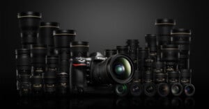 A Nikon DSLR camera with a lens attached is surrounded by multiple Nikon lenses of various sizes, all displayed against a dark background.