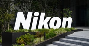 Large, white "Nikon" sign stands amid greenery outside a modern building, near concrete steps and surrounded by trees and bushes.