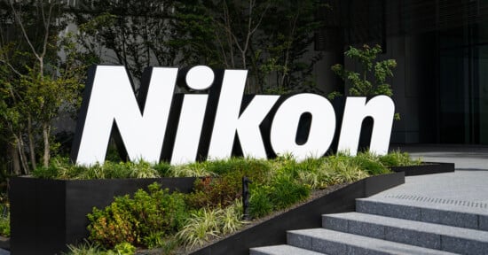 Large, white "Nikon" sign stands amid greenery outside a modern building, near concrete steps and surrounded by trees and bushes.