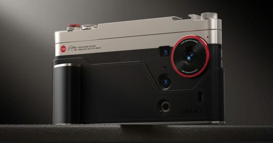 A close-up of a modern camera with a metallic top, black leather texture, and a prominent lens outlined in red. The camera has several buttons, dials, and is resting on a dark surface.