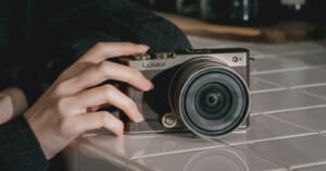 A hand rests on a white tiled surface, holding a Lumix digital camera with a large lens, positioned at an angle for a close-up view.