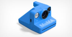 A blue instant camera with the word "MoMA" printed on the front, featuring a prominent lens, viewfinder, and flash against a plain white background.