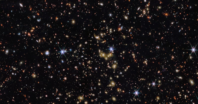 A deep space image showing countless distant galaxies, stars, and bright points of light scattered across a black background, with some stars displaying diffraction spikes.