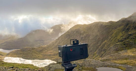 A vintage camera on a tripod faces a misty mountain landscape with rolling hills, rocky terrain, and reflective lakes under a dramatic cloudy sky.