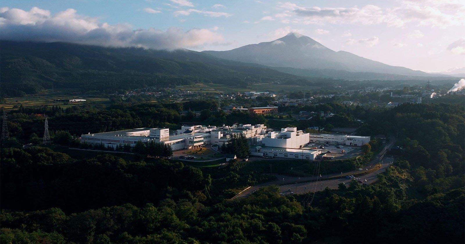 A large white industrial facility surrounded by lush green trees, with a mountain in the background under a partly cloudy sky. Roads and small buildings are visible in the vicinity.