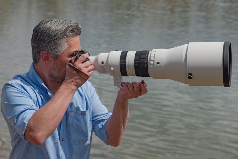 A man with gray hair and a beard, wearing a light blue shirt, is photographing with a large, white telephoto camera lens beside a body of water.