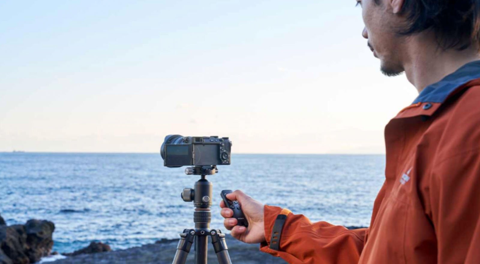 A person in an orange jacket stands by the sea, holding a remote while operating a camera mounted on a tripod, with the ocean and sky in the background.