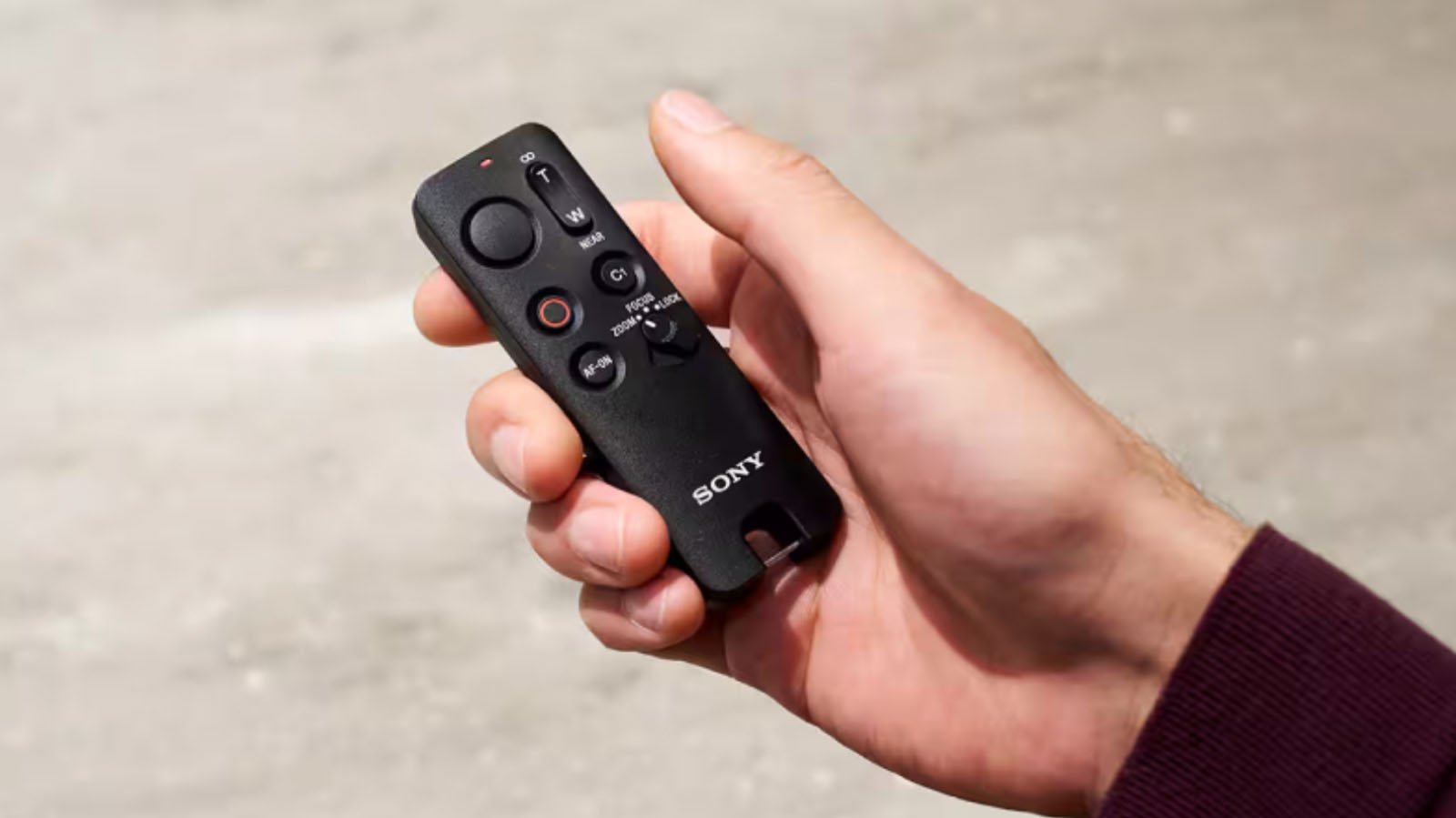A person holding a small black Sony remote control with several buttons and a joystick, against a blurred light gray background.