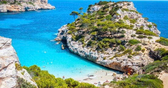 A small, sandy beach with turquoise water is nestled between rocky cliffs covered with green trees and shrubs, with people swimming and sunbathing under a clear blue sky.