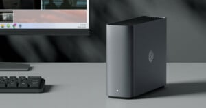 A sleek, dark gray desktop computer tower sits on a desk next to a keyboard and a monitor, featuring a small white LED light and a logo on the front panel.