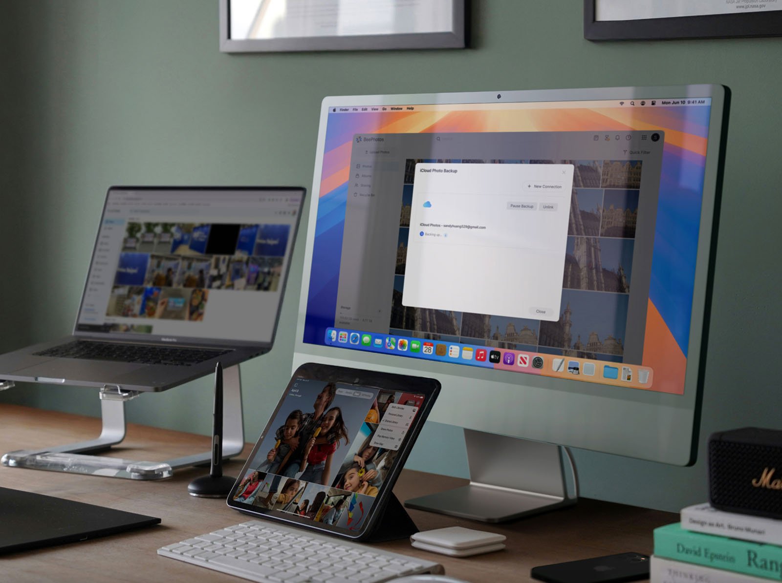 A modern workspace with a desktop computer, laptop, and tablet, all displaying different screens. A white keyboard and mouse are on the wooden desk, which also has stacked books and headphones. The background wall is green.