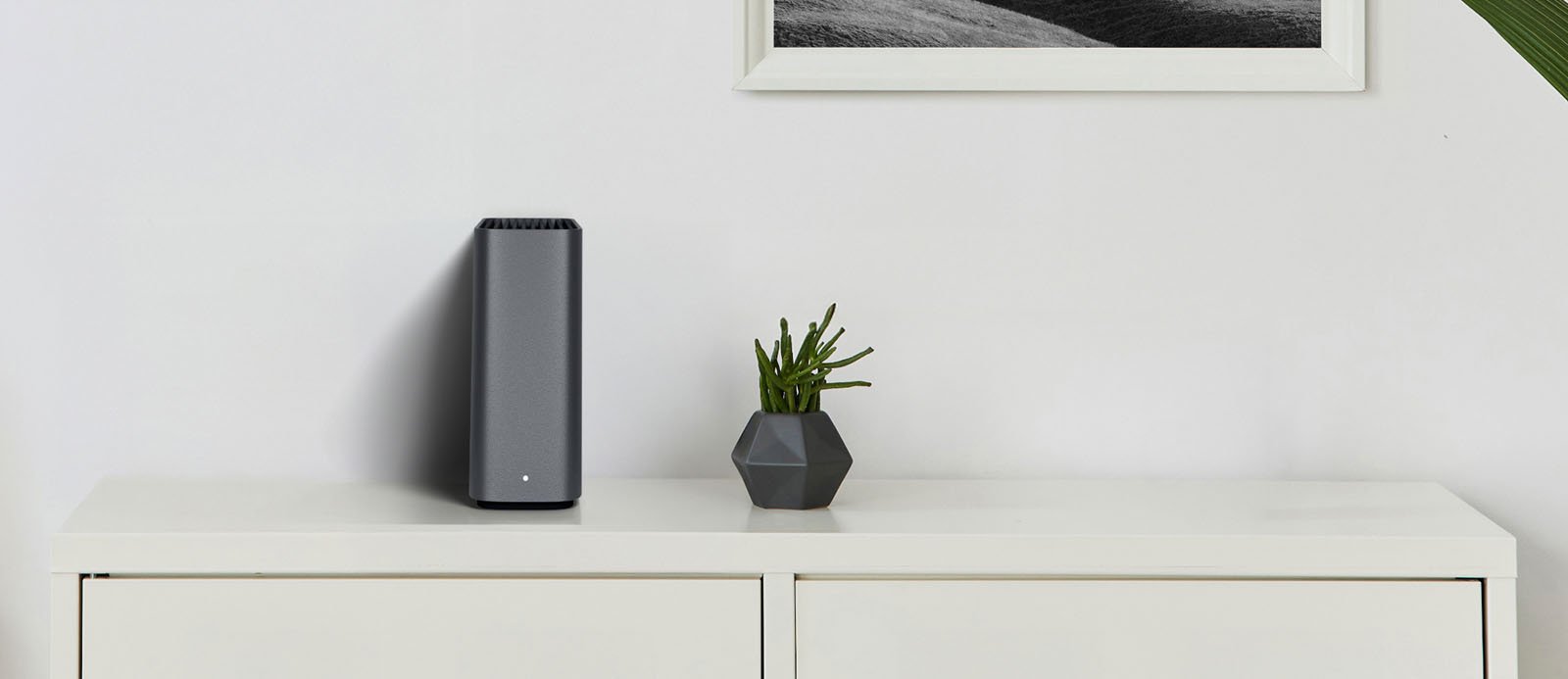 A modern gray electronic device, a small geometric plant pot, and a black-and-white framed photo sit on a white cabinet against a white wall. A green plant leaf is visible on the right edge of the image.