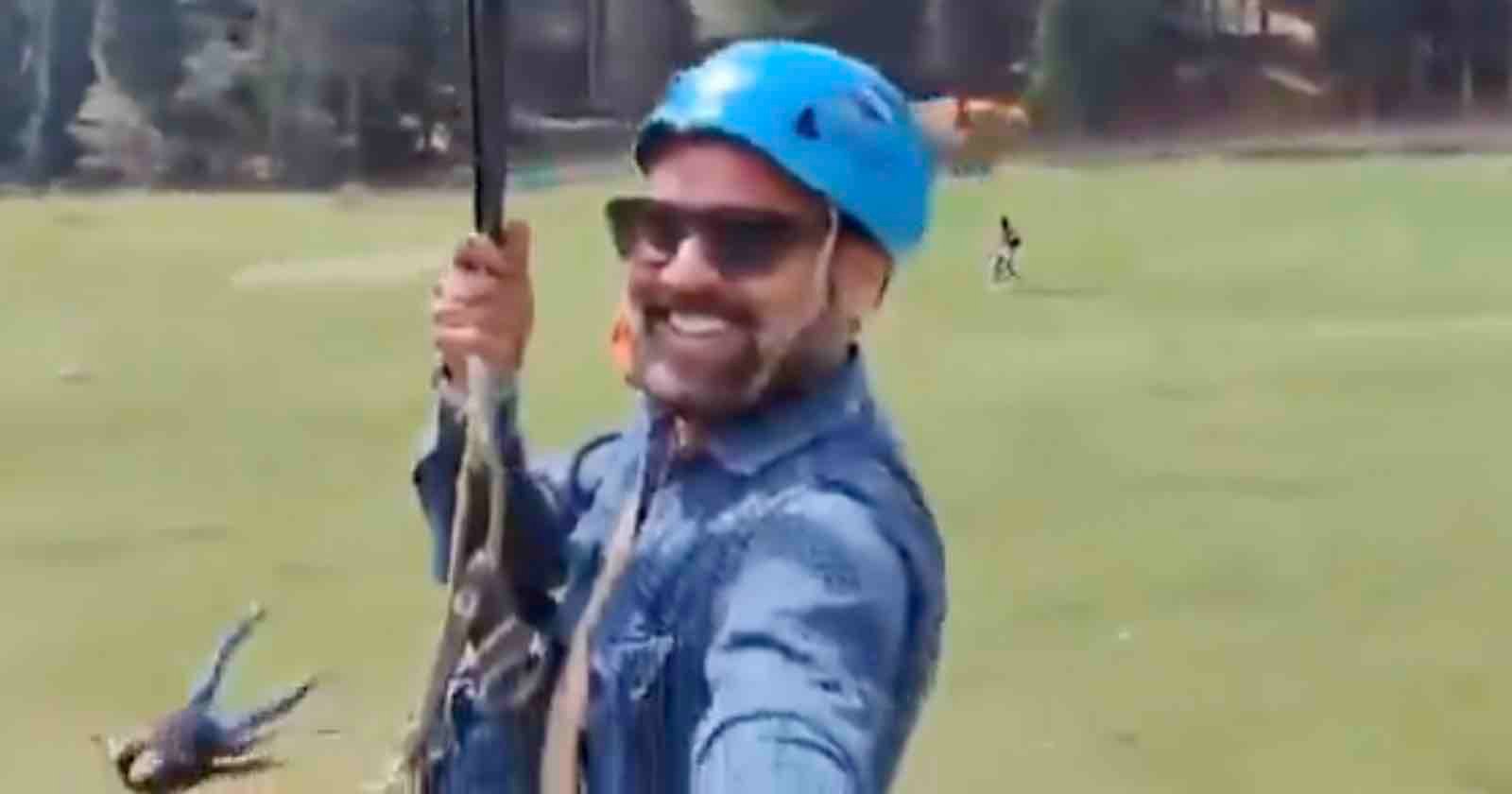 A smiling man wearing sunglasses, a blue helmet, and a denim shirt holds onto a zipline rope while gliding over a grassy field, with trees and other people visible in the background.