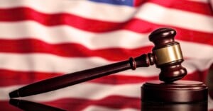 A wooden judge’s gavel rests on a sound block with a blurred American flag in the background.