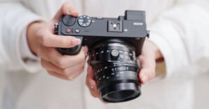 A person in a white jacket holds a black digital camera with both hands, adjusting the lens and dials. The camera and the person’s hands are in sharp focus.