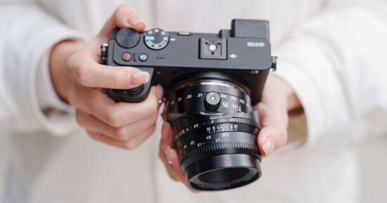 A person in a white jacket holds a black digital camera with both hands, adjusting the lens and dials. The camera and the person’s hands are in sharp focus.