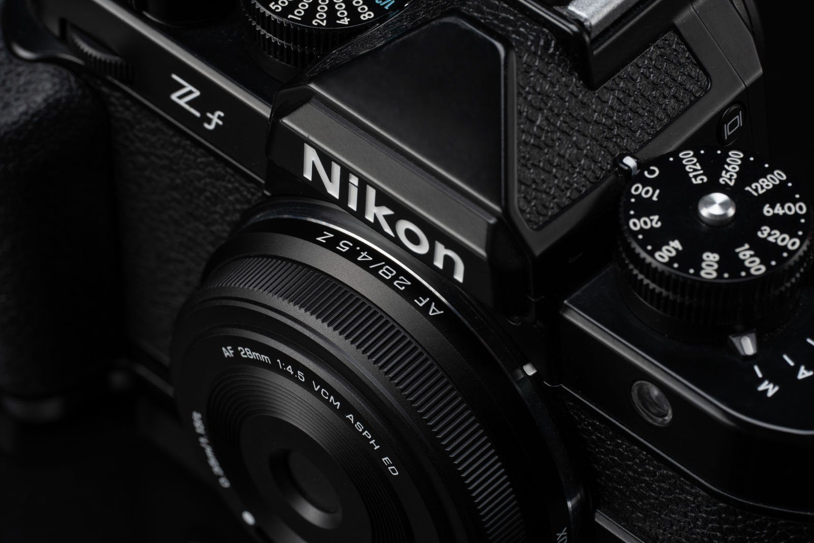 Close-up view of a black Nikon Z f camera, showing the lens with markings, textured grip, and control dials with shutter speed numbers.