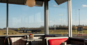 Inside a diner with red booths and a condiment-filled table by the window, overlooking a highway, grassy fields, and a yellow truck passing by under a clear sky.