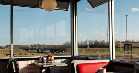 Inside a diner with red booths and a condiment-filled table by the window, overlooking a highway, grassy fields, and a yellow truck passing by under a clear sky.