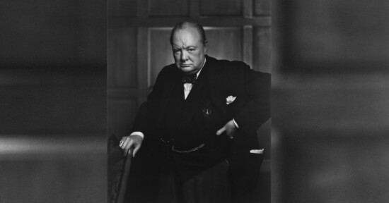 A serious older man in a suit stands with one hand on his hip and the other resting on a chair, in front of a paneled wall. He wears a bow tie and pocket square and looks directly at the camera.