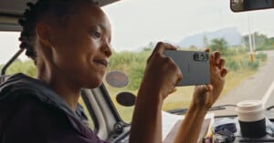 A person with short hair sits in a vehicle, holding a smartphone sideways to take a photo. A travel mug and papers are on the dashboard, and a road with greenery is visible through the window.