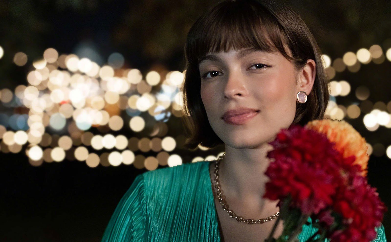A young woman with short brown hair and earrings smiles softly, holding a bouquet of colorful flowers. She wears a green top and gold chain necklace. The background is blurred with glowing lights at night.