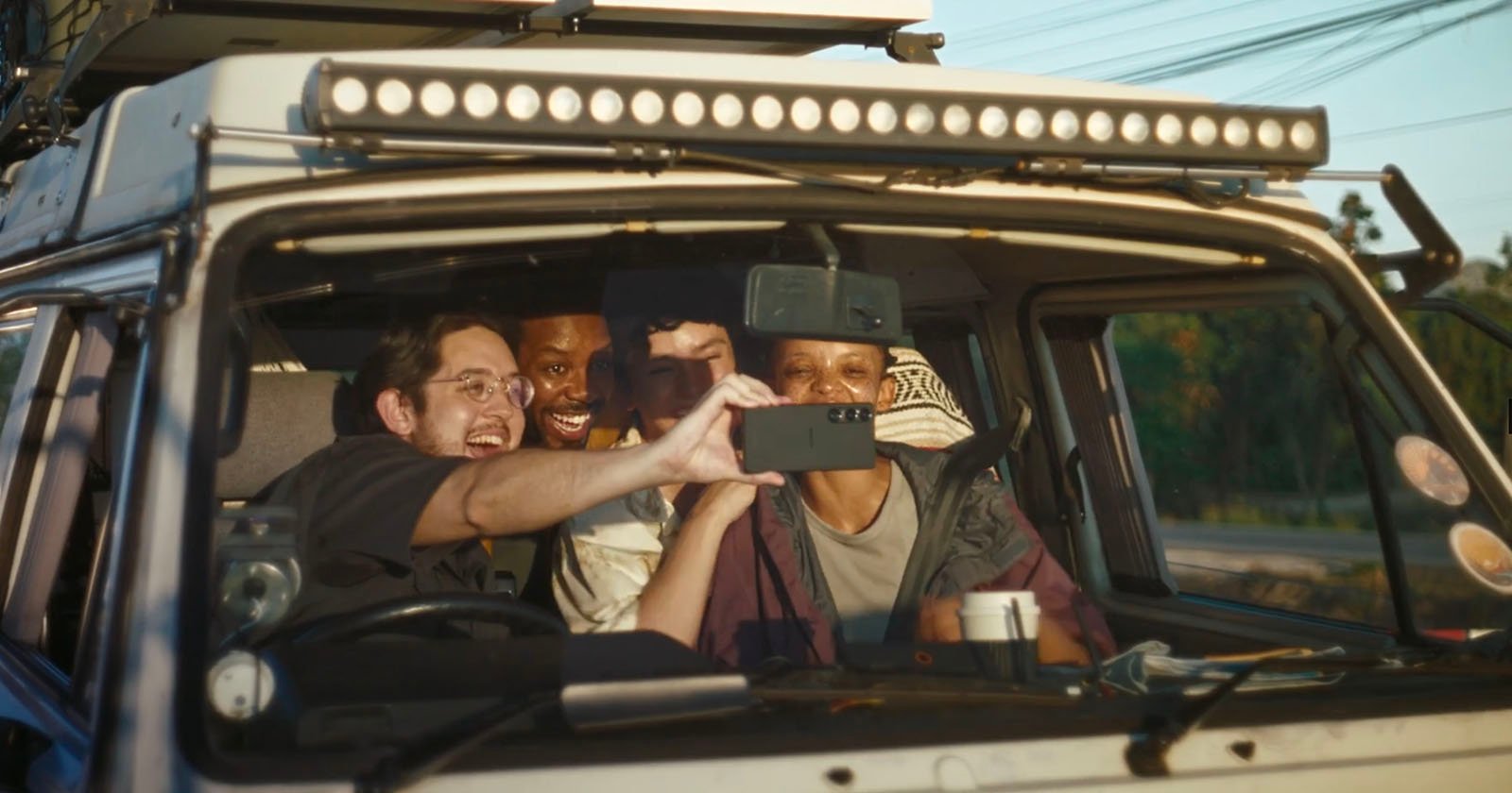 Four friends sit in the front seats of a vehicle, smiling and crowded together as they take a selfie with a smartphone. Sunlight streams through the windows, creating a cheerful and adventurous atmosphere.