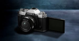 A Yashica FX-D camera with a black and silver body, featuring a Yashica lens and a flip-out LCD screen, is placed on a dark blue textured surface.