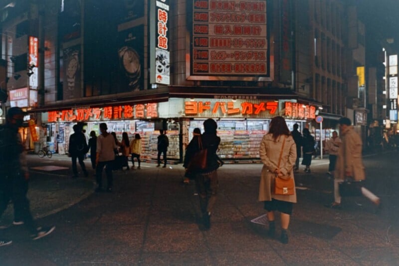 A busy city street at night with people walking; bright neon signs in Japanese illuminate a corner electronics store. Two people stand in the foreground facing the shop.