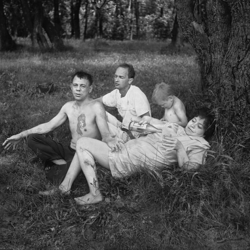 Four adults relax in the grass near a tree in a wooded area. One man is shirtless with visible tattoos; a woman reclines and smiles while reading a magazine, and another man and woman sit nearby, enjoying the outdoors.