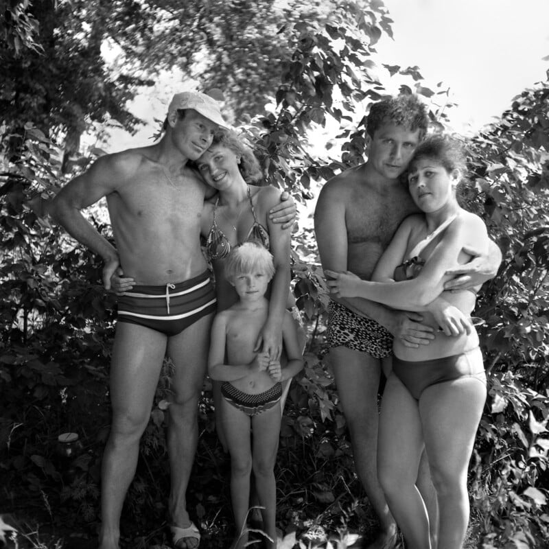 A black and white photo of two men and two women in swimsuits standing closely together beside a young child outdoors, surrounded by leafy bushes and trees, all looking at the camera.