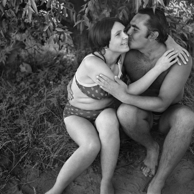 A man and woman in swimsuits sit on the ground outdoors, embracing and smiling affectionately under the shade of leafy trees. The scene is relaxed and intimate, suggesting a warm, sunny day.