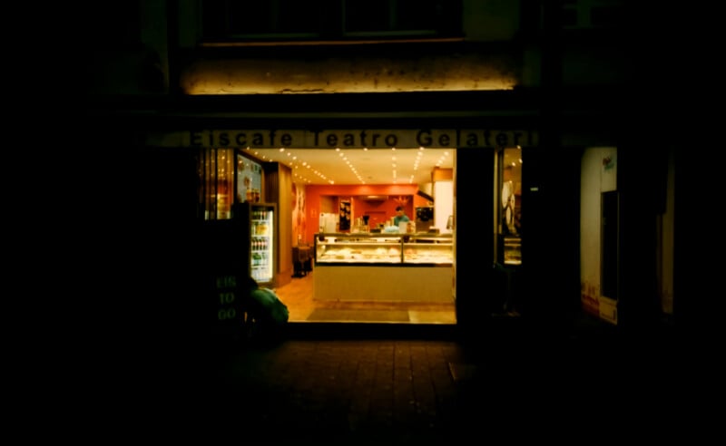 A warmly lit gelato shop with large glass doors open to the street at night. Inside, display cases show various gelato flavors, and the shop interior features bright lights and a welcoming atmosphere.