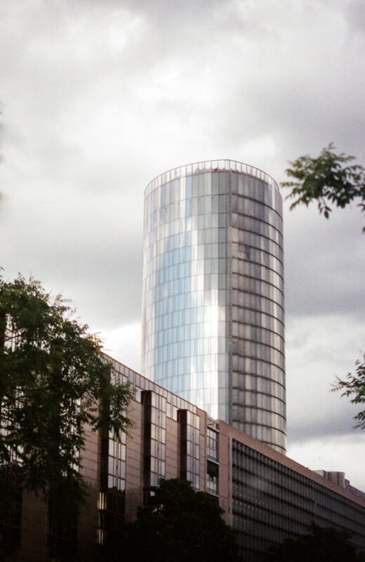 A tall, cylindrical glass skyscraper reflects the cloudy sky, standing behind a lower, modern building with trees partially framing the scene.