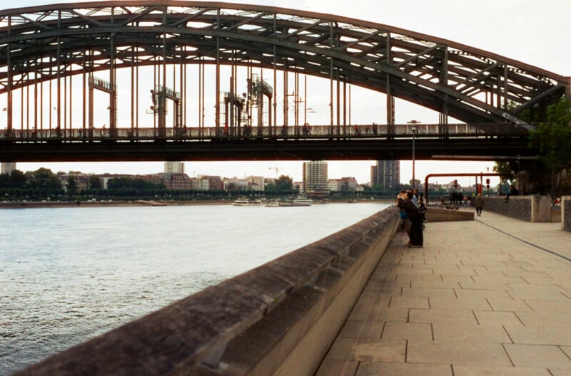 A wide river runs alongside a paved riverside walkway, with people walking and leaning on the railing. An arched steel bridge spans the river, and city buildings are visible in the background.