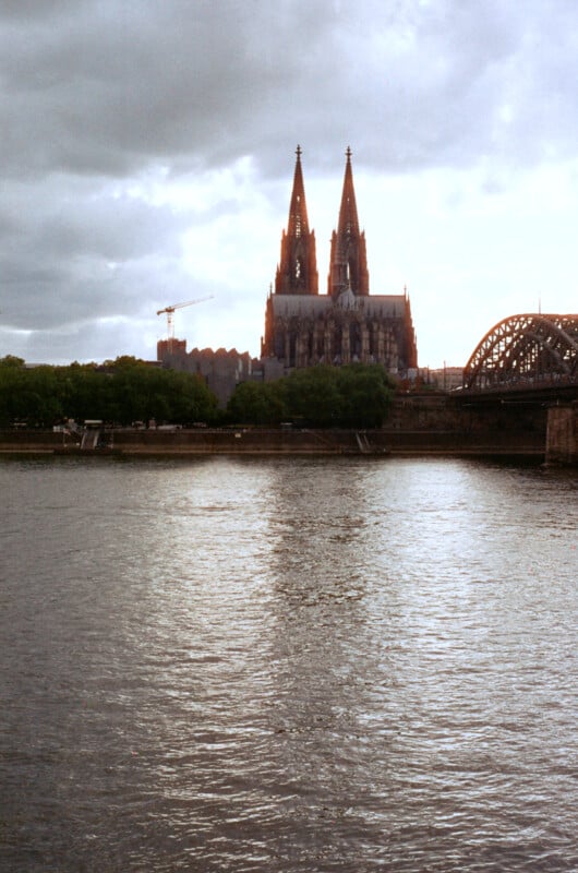 Cologne Cathedral with its twin spires rises above trees and buildings near the Rhine River, under a cloudy sky. The Hohenzollern Bridge is visible on the right.