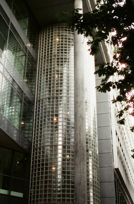 A modern building facade featuring a tall cylindrical section made of glass blocks, reflecting light, with tree branches partially visible in the upper right corner.