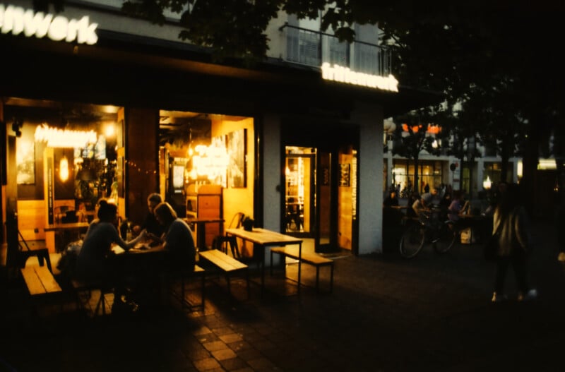 A warmly lit café at night with people sitting at wooden tables outside under trees, talking and eating. The building glows with yellow light, and "innwerk" is visible on the sign above the entrance.