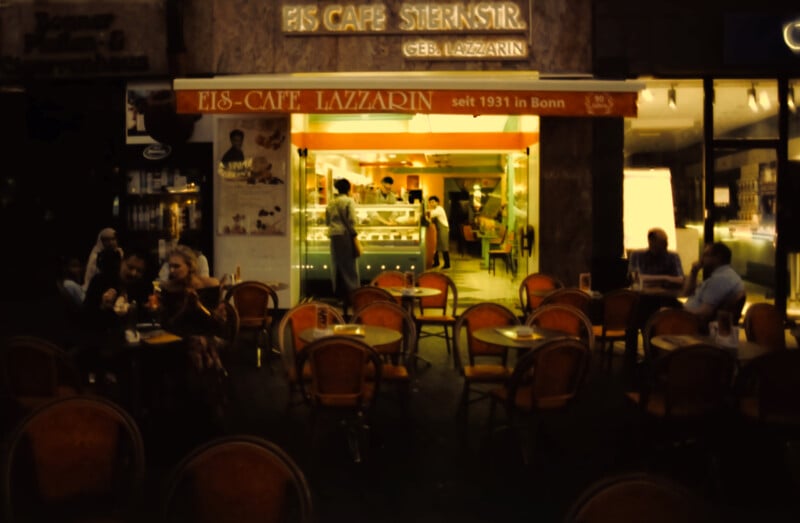 A warmly lit ice cream café called "Eis-Café Lazzarin" with empty and occupied outdoor tables at night, and people visible inside near a counter. The sign indicates it has been in Bonn since 1931.